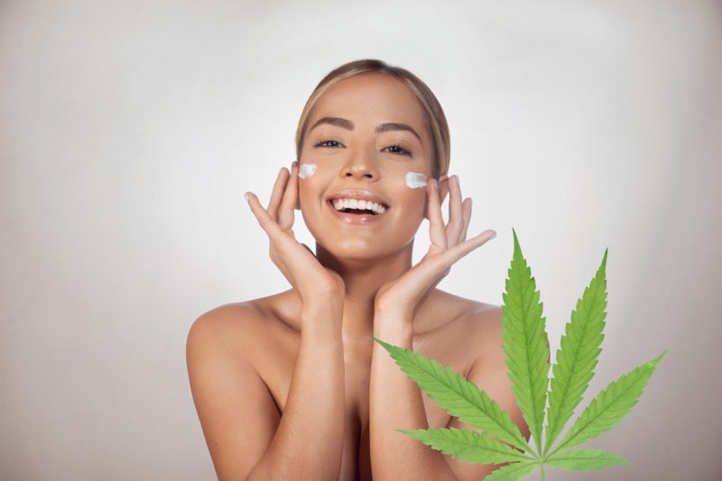 Brunette woman applying CBD facial cream made from cannabis extract for a natural cbd skincare treatment. Portrait of young woman with cannabis leaf. Cosmetology and treatment concept. Isolated on gray background